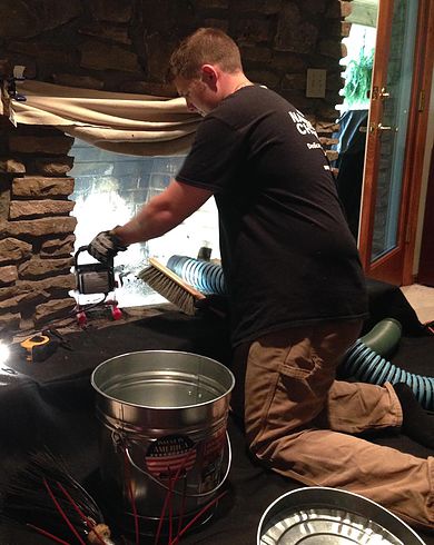 Traditions Chimney Sweep original photo of tech cleaning firebox. Tech cleaning stack rock fireplace with broom and vacuum and bucket in front.
