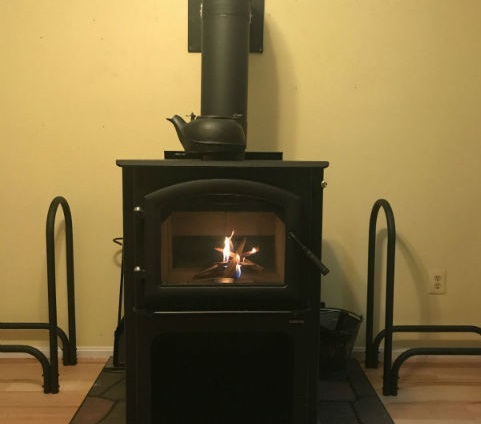 Black freestanding wood stove with racks for wood on each side and cast iron pot sitting on top.
